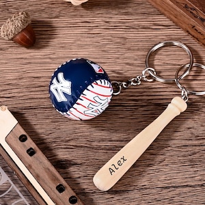 Könnte beinhalten: Ein Schlüsselanhänger mit einem Miniatur-Baseball mit blau-weißem New York Yankees-Logo und einem kleinen hölzernen Baseballschläger-Anhänger, in den der Name Alex eingraviert ist. Beide Teile sind an einem silbernen Metallring befestigt.
