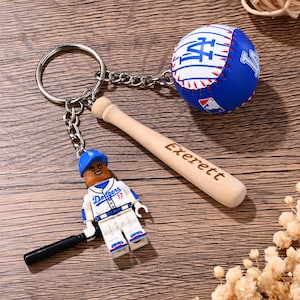 Op de afbeelding: Een sleutelhanger met een metalen ring en ketting waaraan een blauw-witte honkbal met het LA-logo, een kleine honkbalknuppel van licht hout met de inscriptie Exerett en een klein plastic figuurtje in een Dodgers-tenue hangen.
