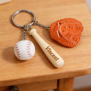 May include: A baseball-themed keychain featuring a white baseball with red stitching, an orange baseball glove, and a wooden bat engraved with "Vincent." The keychain is attached to a silver keyring.