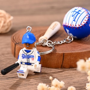 Op de afbeelding: Een Los Angeles Dodgers honkbal-sleutelhanger met een minifiguur in een wit en blauw uniform met nummer 17, een blauwe pet, een zwarte knuppel en een blauw-witte honkbalbedel aan een zilverkleurige metalen ring en ketting.