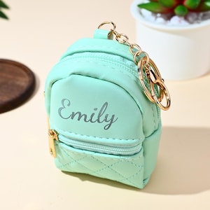 Peut inclure: Un porte-clés mini sac à dos vert menthe avec une fermeture éclair et des accessoires dorés. Le mot "Emily" est imprimé sur le devant. Le petit sac a un motif matelassé en bas et une chaîne dorée.