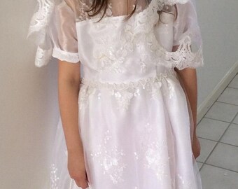 Puede incluir: Una niña pequeña con un vestido blanco de encaje, velo y corona de flores. Lleva un pequeño bolso blanco y tiene el pelo recogido.