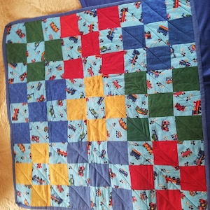 May include: A patchwork quilt with a checkered pattern in blue, green, yellow, and red. The quilt is made of fabric with a small print of toy trucks.