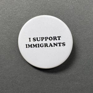 May include: A white, circular button with the text "I SUPPORT IMMIGRANTS" in bold, black letters. The button is centered on a gray background.