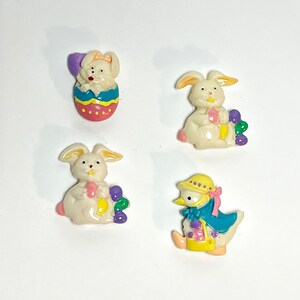 May include: Four Easter-themed ceramic figurines: a white bunny holding a purple egg, a white bunny in an egg-shaped basket, a white bunny holding a purple flower, and a duck wearing a yellow hat and a blue coat.