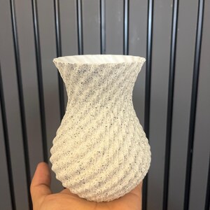 May include: A textured, off-white vase with a spiral design. The vase has a wide, flared top and a rounded base. The surface is speckled with dark gray flecks, adding visual interest. The vase is held in a hand, showcasing its size and shape.