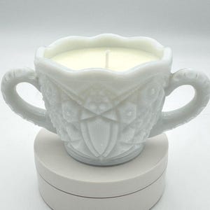 Puede incluir: Una vela blanca en una taza decorativa de estilo vintage con dos asas. La taza tiene un diseño texturizado con forma de diamante. La cera de la vela es de color blanco crema, y se ve una sola mecha en el centro. La taza se apoya sobre una base redonda blanca.