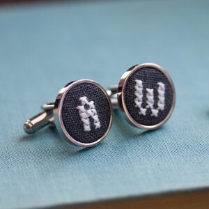 May include: A pair of silver cufflinks with a dark gray fabric face. Each cufflink features a white embroidered letter, one with an "A" and the other with a "W". The cufflinks have a classic, round design.