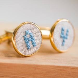 May include: A pair of gold cufflinks with a white fabric background and blue embroidered letters. The letters are "A" and "Y".