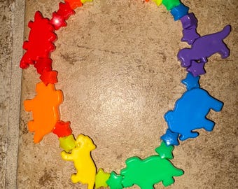 Puede incluir: Un collar colorido con cuentas en forma de estrella y colgantes de animales. Los colgantes incluyen un dinosaurio rojo, un oso naranja, un perro amarillo, un dinosaurio verde, un oso azul y un perro morado.