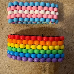 May include: Two beaded bracelets. The top bracelet features a design with light blue, pink, and white beads. The bottom bracelet displays a rainbow pattern with red, orange, yellow, green, blue, and purple beads. Both bracelets are made of small, round beads.