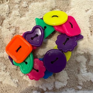 May include: A colorful bracelet made of various shaped buttons. The bracelet features orange squares, purple hearts, green triangles, pink rectangles, yellow circles, and purple flower-shaped buttons. The buttons are connected by a black string.