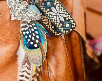 May include: A decorative charm featuring a silver butterfly, a blue gemstone, and colorful feathers. The charm also includes a patterned fabric band with floral designs. The background is a brown leather surface.