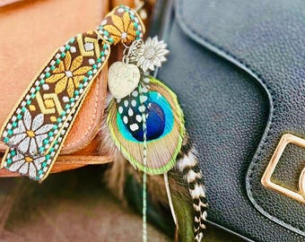 May include: A decorative bag charm featuring a colorful peacock feather, a heart-shaped charm, and a sunflower charm. The charm also includes a patterned tassel with yellow and white floral designs, attached to a gold chain.