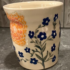 May include: A ceramic mug with a cream-colored base. The mug features a large orange floral design on one side and blue floral sprigs on the other. The flowers are hand-painted with a delicate touch, adding a touch of artistry to the piece.