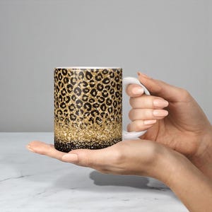 May include: A ceramic mug featuring a black and gold leopard print pattern that transitions into a dense gold glitter effect at the base, finished with a solid white handle and a smooth, glossy surface texture throughout the exterior.