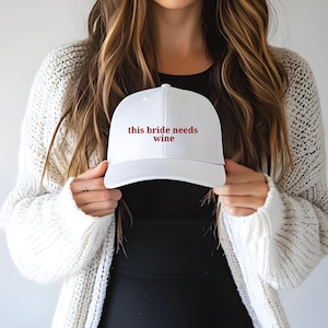 May include: White baseball cap with the text "this bride needs wine" in burgundy lettering. The cap is held up by a person wearing a black top and a white cardigan.