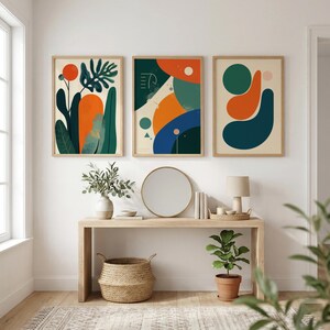 Könnte beinhalten: Drei gerahmte abstrakte Kunstdrucke mit botanischen und geometrischen Designs in Orange, Grün und Blau. Die Drucke werden über einer Holzkonsolentisch mit einem Spiegel, einer Vase und dekorativen Gegenständen ausgestellt.