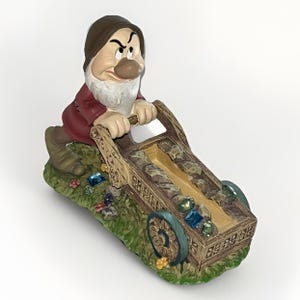Peut inclure: Une figurine de nain Grincheux portant un bonnet marron et une tunique rouge, poussant une brouette en bois remplie de fausses pierres précieuses colorées. La base est texturée comme de l'herbe verte avec des fleurs.