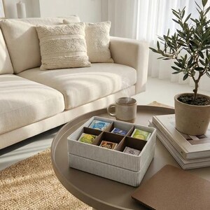 May include: A living room scene with a beige sofa, decorative pillows, and a round coffee table. A box of tea bags sits on the table, alongside a mug, a small potted plant, and books. The room is bathed in natural light.