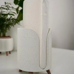 May include: A white paper towel holder with a textured, ribbed design. The holder has a cylindrical shape with a slot for dispensing towels. It is elevated on three wooden legs, and a roll of white paper towels is inside.