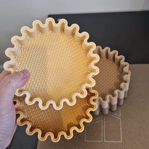 May include: Three beige, round, decorative trays with a wavy, scalloped edge. The trays have a textured interior surface. One tray is being held by a hand. The other two trays are stacked on top of each other.