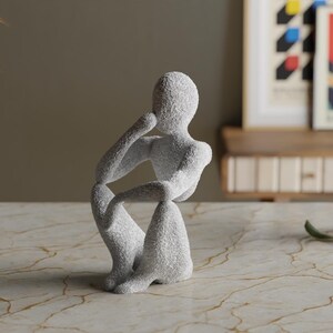 Puede incluir: Una escultura gris texturizada de una persona en una pose contemplativa sentada. La figura descansa la barbilla sobre una mano, con la otra mano en la rodilla. La escultura est&aacute; sobre una superficie con vetas de m&aacute;rmol.
