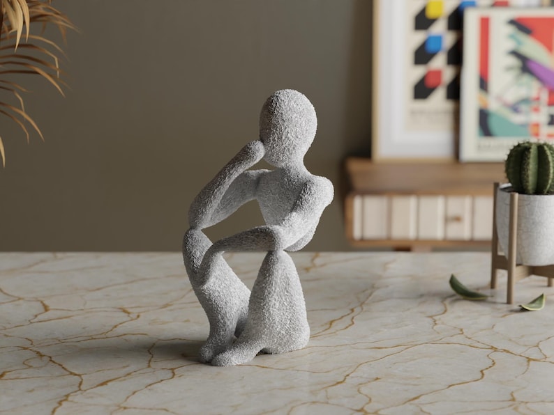 Puede incluir: Una escultura gris texturizada de una persona en una pose contemplativa sentada. La figura descansa la barbilla sobre una mano, con la otra mano en la rodilla. La escultura est&aacute; sobre una superficie con vetas de m&aacute;rmol.