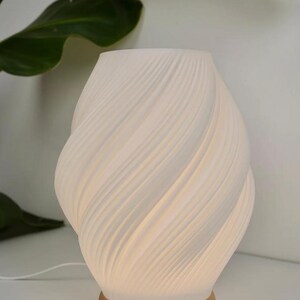 May include: A white ceramic table lamp with a swirling, textured design. The lamp has a warm, soft glow and a tan base. The lamp is near a green plant.