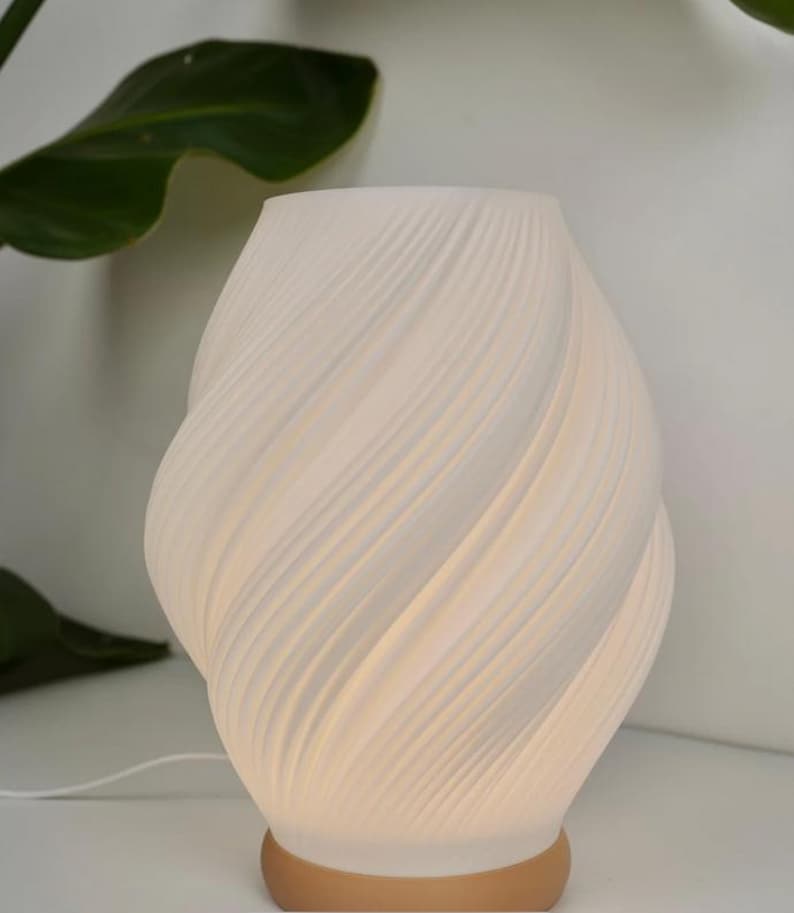 May include: A white ceramic table lamp with a swirling, textured design. The lamp has a warm, soft glow and a tan base. The lamp is near a green plant.