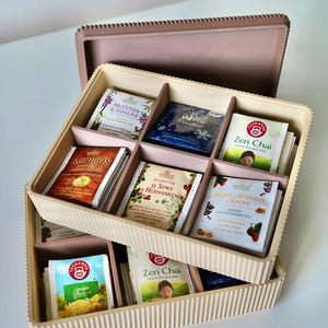 May include: A beige tea box with a lid, filled with various tea bags. The tea bags are in different colors and designs, with text labels. The box is divided into compartments, and the lid is open, revealing the tea bags inside.