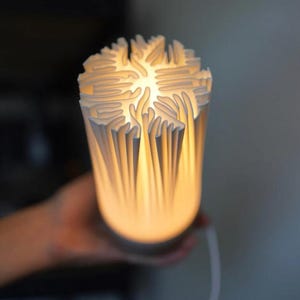 Puede incluir: Una l&aacute;mpara de cer&aacute;mica blanca con un dise&ntilde;o texturizado &uacute;nico. La parte superior de la l&aacute;mpara presenta un patr&oacute;n detallado y en capas, y el interior brilla con una luz c&aacute;lida y suave. La l&aacute;mpara es cil&iacute;ndrica y tiene una est&eacute;tica moderna.