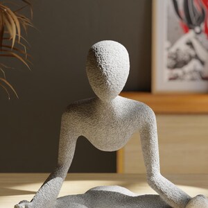 May include: A light gray, textured sculpture of a person in a seated yoga pose. The figure has a smooth, rounded head and arms extended to the sides. The sculpture is on a wooden surface.