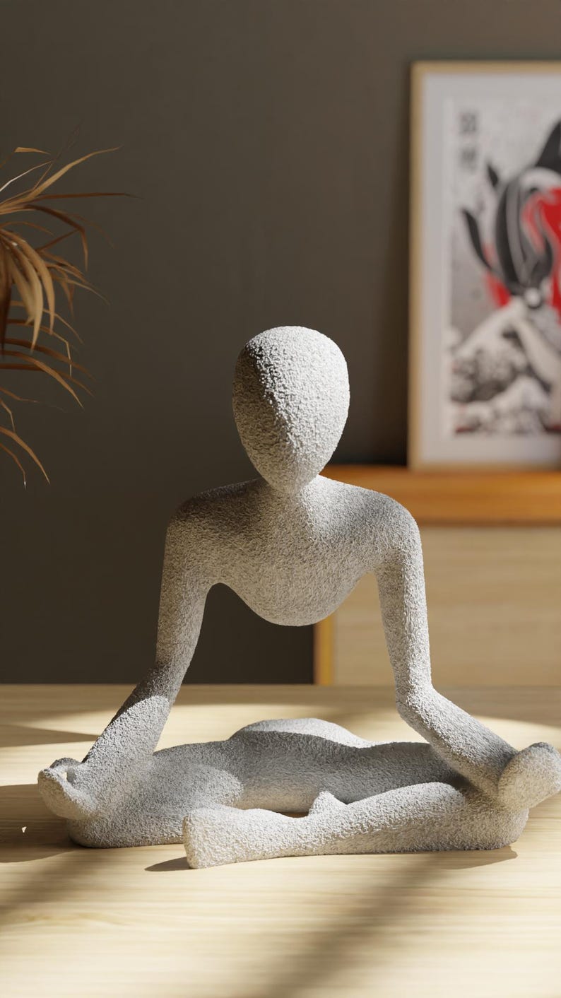 May include: A light gray, textured sculpture of a person in a seated yoga pose. The figure has a smooth, rounded head and arms extended to the sides. The sculpture is on a wooden surface.