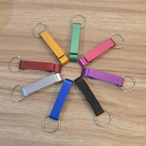 May include: A selection of bottle opener keyrings in a circular arrangement. The keyrings are in a variety of colours: gold, green, pink, purple, black, blue, red, and silver. Each keyring has a metal ring attached.
