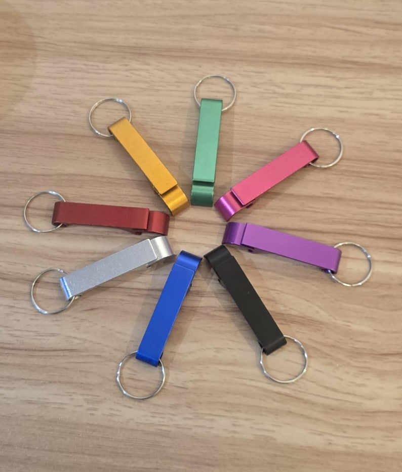 May include: A selection of bottle opener keyrings in a circular arrangement. The keyrings are in a variety of colours: gold, green, pink, purple, black, blue, red, and silver. Each keyring has a metal ring attached.