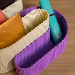May include: A desk organiser featuring two curved, flexible compartments with a textured, scalloped edge. One compartment is light beige, and the other is purple. A turquoise highlighter and a gold envelope are tucked inside the organiser.