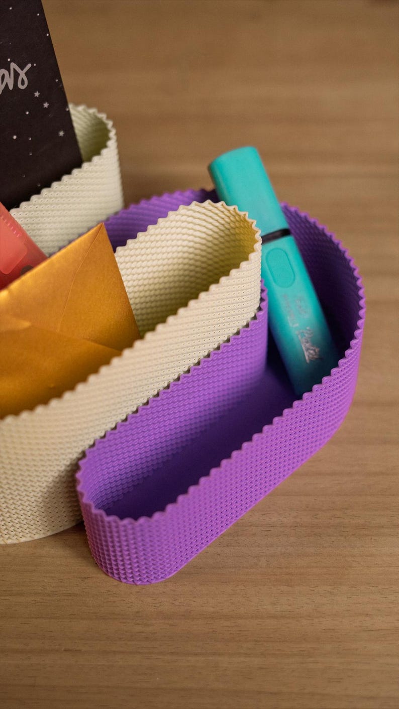 May include: A desk organiser featuring two curved, flexible compartments with a textured, scalloped edge. One compartment is light beige, and the other is purple. A turquoise highlighter and a gold envelope are tucked inside the organiser.