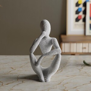 Puede incluir: Una escultura texturizada, de color gris claro, de una persona en una pose sentada y pensativa. La figura tiene la barbilla apoyada en una mano y las piernas cruzadas. La escultura est&aacute; sobre una superficie similar al m&aacute;rmol.