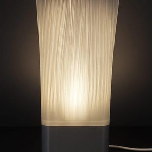 May include: A modern table lamp with a rectangular gray base and a translucent, textured shade. The shade is lit from within, creating a soft, warm glow. A white power cord extends from the base.