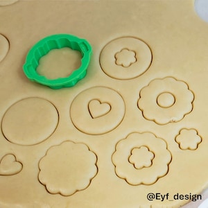 May include: A bright green plastic cookie cutter rests on a sheet of rolled-out beige dough. The dough features various cut-out shapes including scalloped circles, plain circles, small hearts, and flower-like forms. The text @Eyf_design is visible.