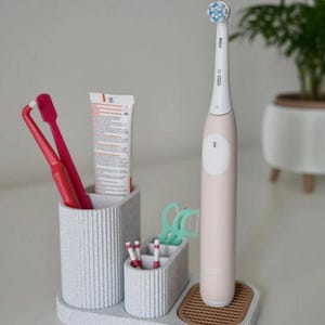 May include: A light gray toothbrush holder with multiple compartments. It holds a pink electric toothbrush, red toothbrushes, toothpaste, floss picks, and small toothbrushes. The holder sits on a tray with a brown, textured base.