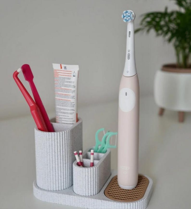 May include: A light gray toothbrush holder with multiple compartments. It holds a pink electric toothbrush, red toothbrushes, toothpaste, floss picks, and small toothbrushes. The holder sits on a tray with a brown, textured base.