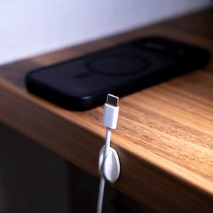 May include: A white cable organiser clip attached to the edge of a wooden surface, holding a white USB-C charging cable in place. A black smartphone with a circular magnetic charging ring on its back is resting on the surface behind the cable.