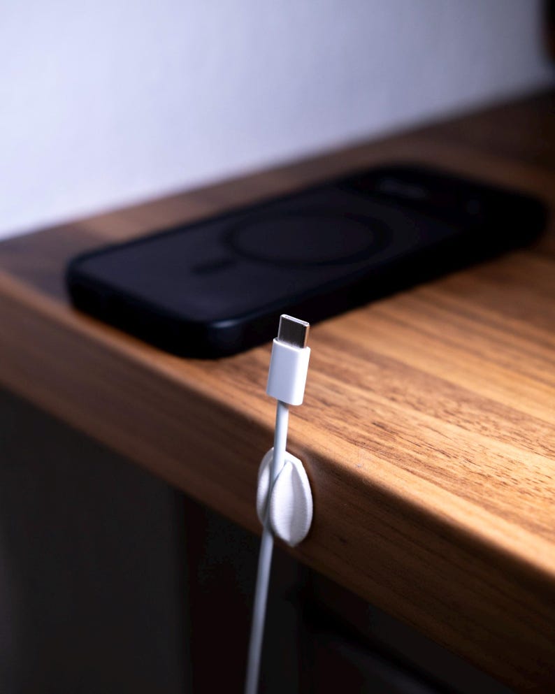 May include: A white cable organiser clip attached to the edge of a wooden surface, holding a white USB-C charging cable in place. A black smartphone with a circular magnetic charging ring on its back is resting on the surface behind the cable.