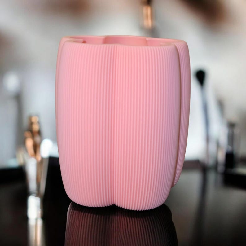 May include: A pink, ribbed, cylindrical container with a tulip-like shape. The container has vertical ridges and a flared top. The object is set against a blurred background, with a dark, reflective surface below.