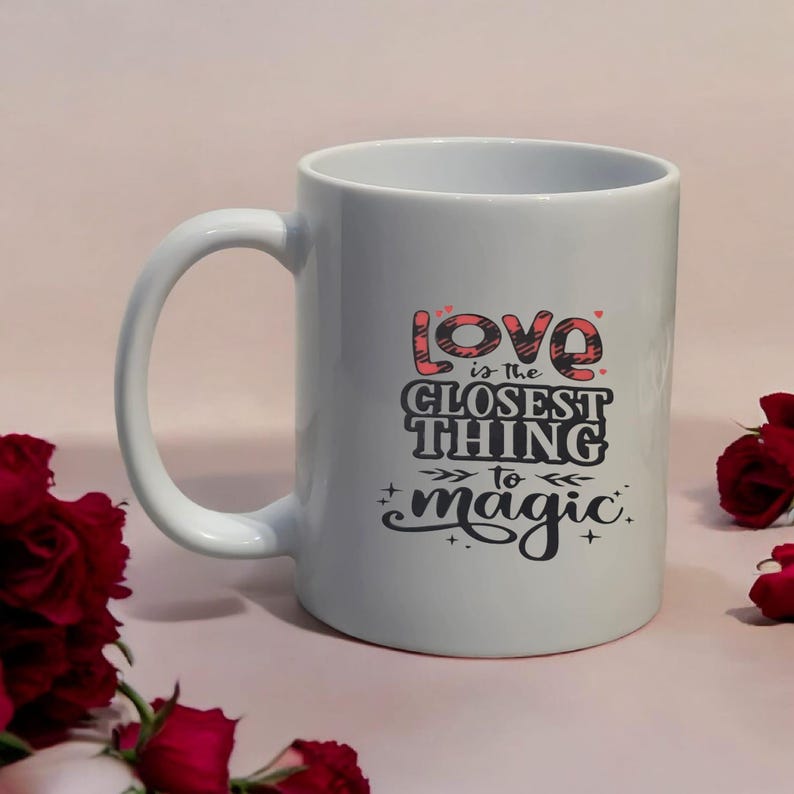May include: White ceramic mug with the text "Love is the closest thing to magic" in a black and red design. The mug has a curved handle and is surrounded by red roses.