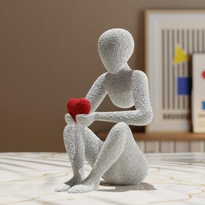 Puede incluir: Una escultura gris texturizada de una figura sentada sosteniendo un coraz&oacute;n rojo. La figura est&aacute; en una pose contemplativa, con el coraz&oacute;n sostenido en sus manos. La escultura est&aacute; sobre una superficie clara.