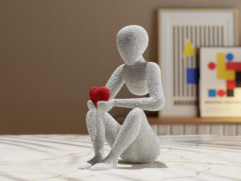 Puede incluir: Una escultura gris texturizada de una figura sentada sosteniendo un coraz&oacute;n rojo. La figura est&aacute; en una pose contemplativa, con el coraz&oacute;n sostenido en sus manos. La escultura est&aacute; sobre una superficie clara.