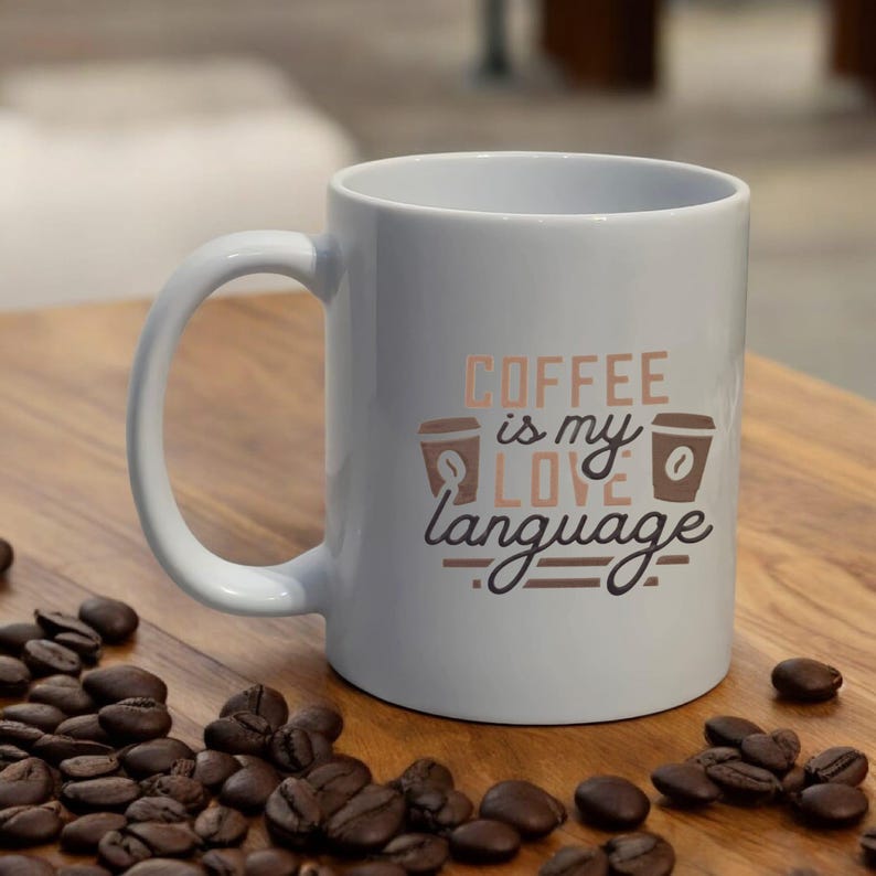 Puede incluir: Taza de caf&eacute; de cer&aacute;mica blanca con el texto "COFFEE is my LOVE language" en letras marrones y cobrizas. La taza presenta dos ilustraciones de tazas de caf&eacute;. Los granos de caf&eacute; est&aacute;n esparcidos alrededor de la base de la taza.